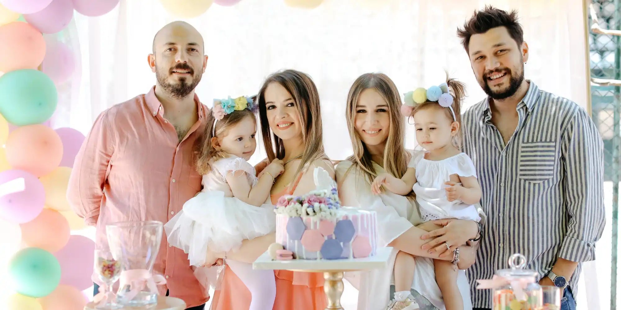 A family of two couples and their two young daughters, celebrating with a pastel-colored birthday cake.