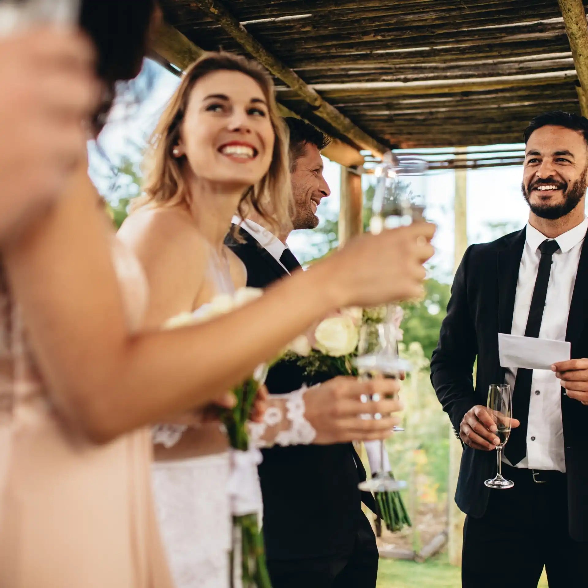 Four people at a celebration, two holding glasses of champagne and two holding bouquets of white roses.