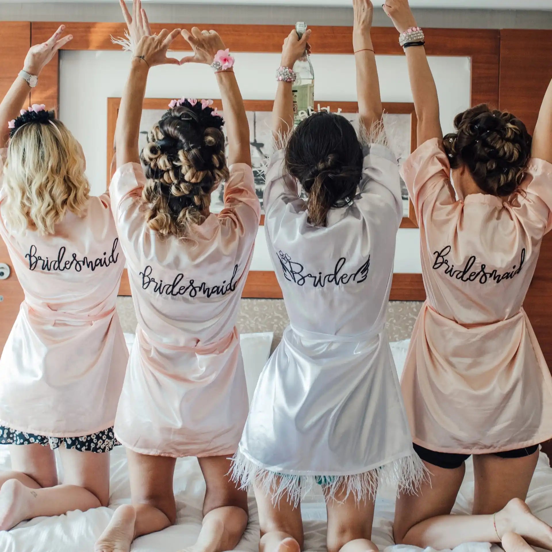 Four women wearing satin robes, three light pink and one white, with ’Bridesmaid’ or ’Bride’ embroidered on the back.