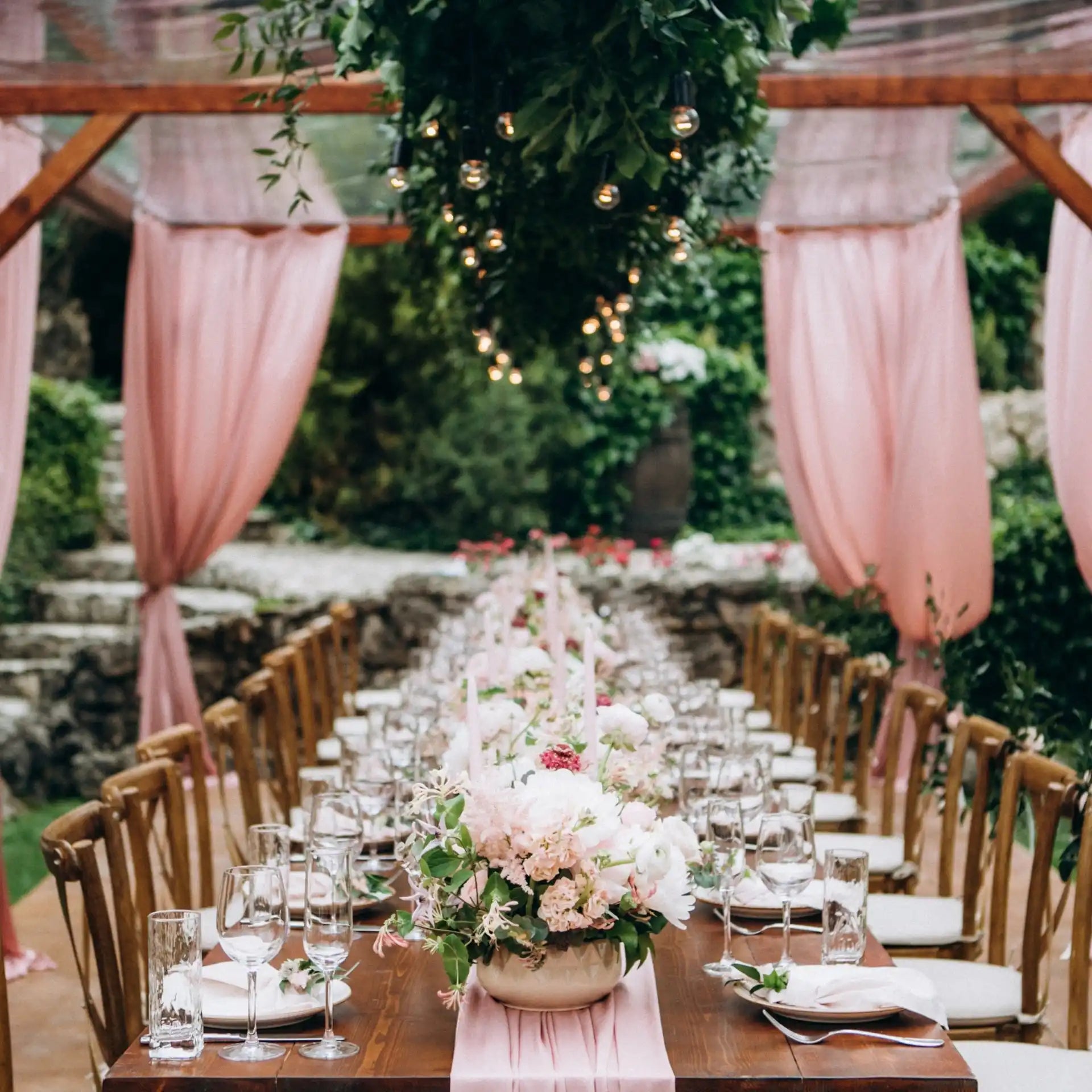 A long wooden table set for an outdoor event with pink floral arrangements, place settings, and chairs.