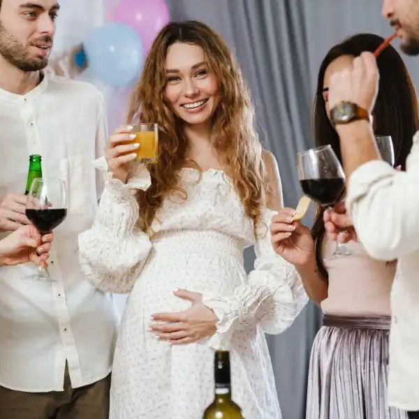 A pregnant woman in a white dress holding a drink, surrounded by friends holding alcoholic beverages.