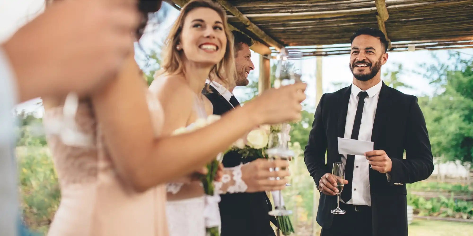 Three people at a celebratory outdoor event, with two holding glasses of champagne, and one holding a note for a speech.