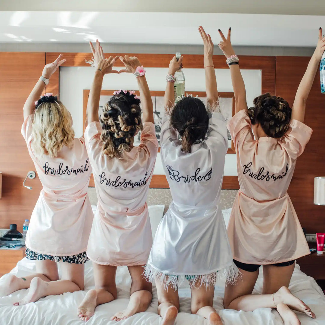 Four people wearing matching satin robes with "Bridesmaid" text kneeling with raised arms on a bed