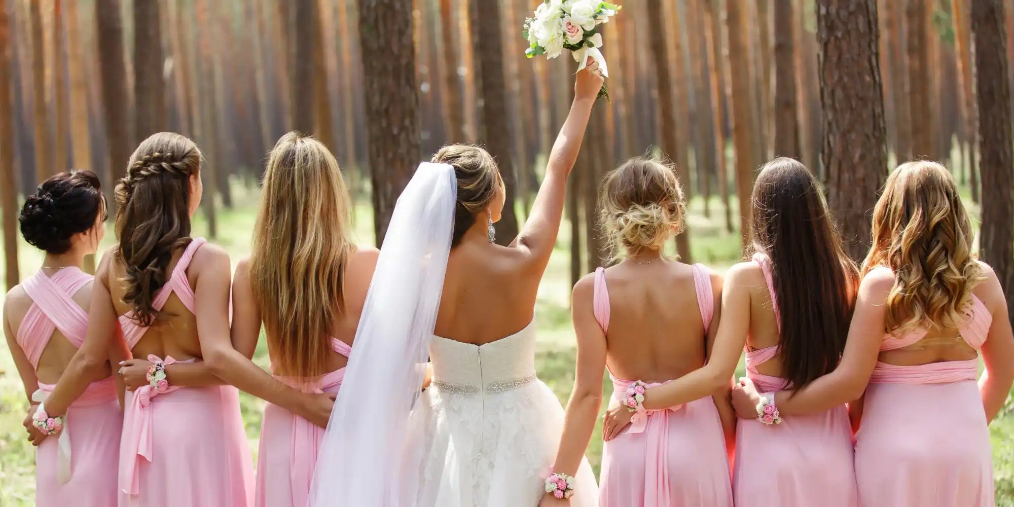 A group of women in formal attire, including a bride and bridesmaids, standing with their backs to the camera in a forest setting.